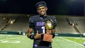 "High school football player in Eagles uniform holding championship trophy on field at night."