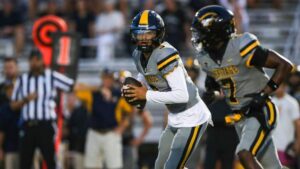 Heritage High QB executing a play during a night football game, closely followed by teammate number seven.